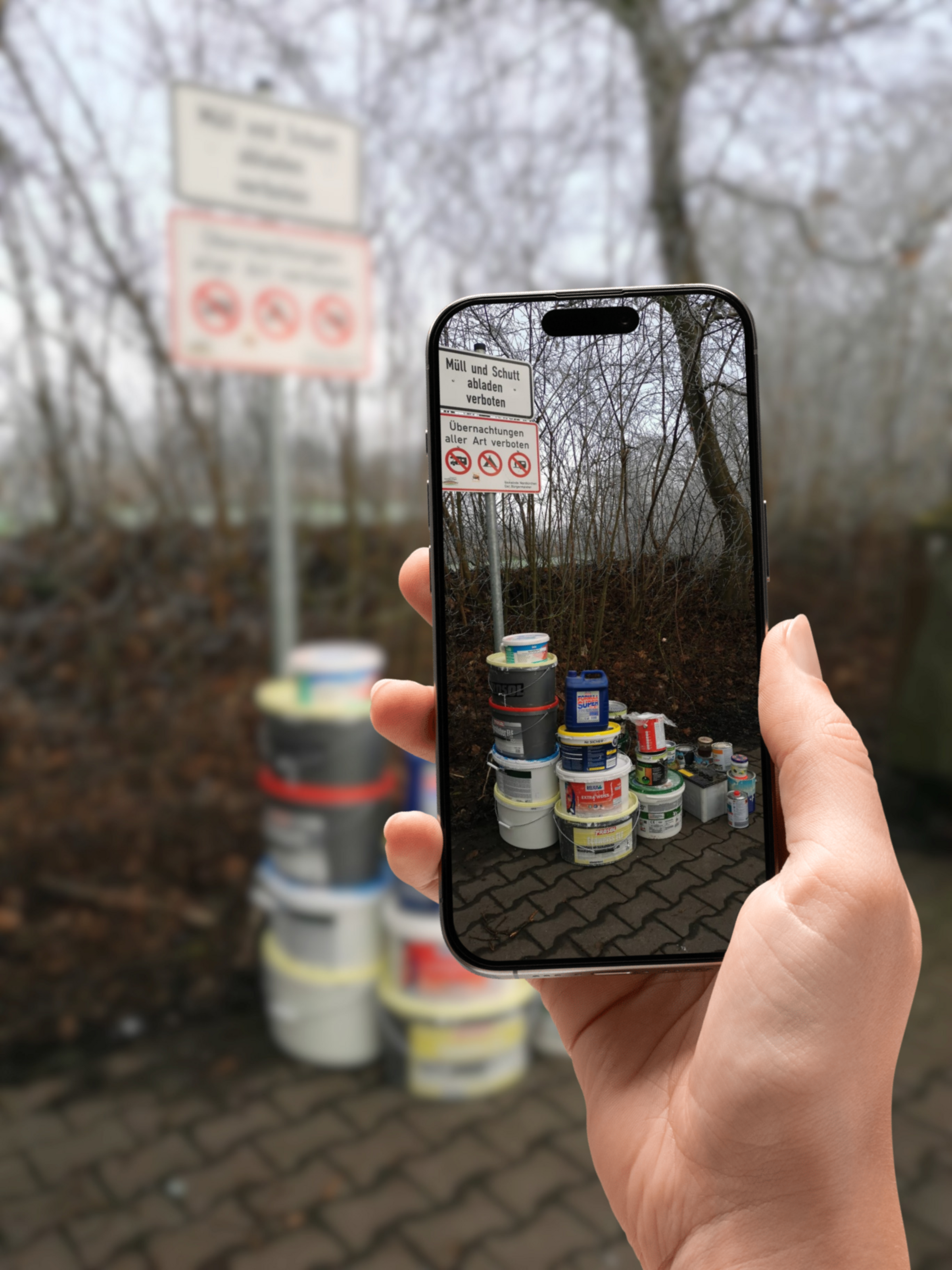 Im Vordergrund eine Hand, die ein Smartphone hält, mit dem eine wilde Müllkippe fotografiert wird